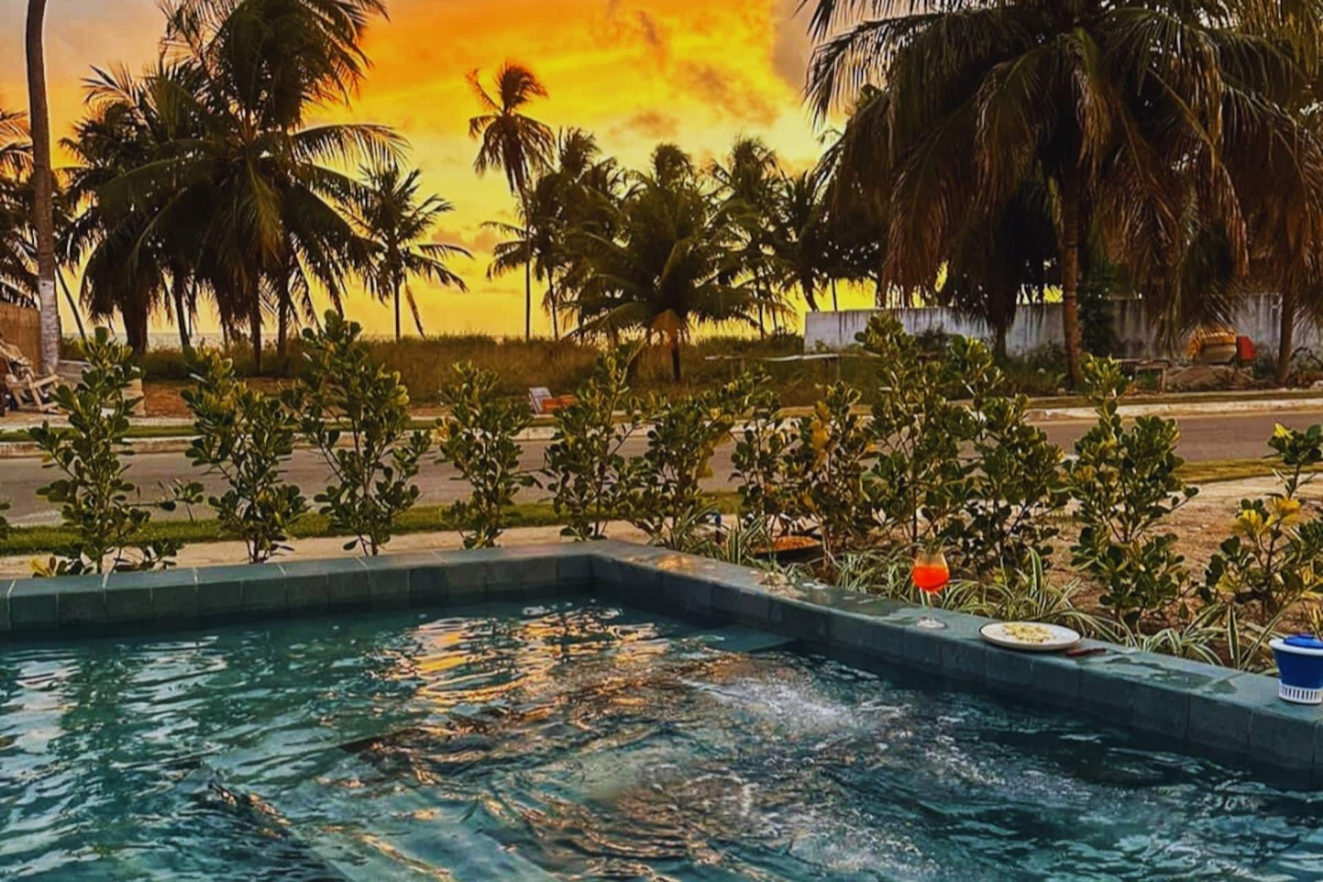 Piscina e hidromassagem da Casa Pérola ao pôr do sol, com coqueiros recortados contra o céu alaranjado