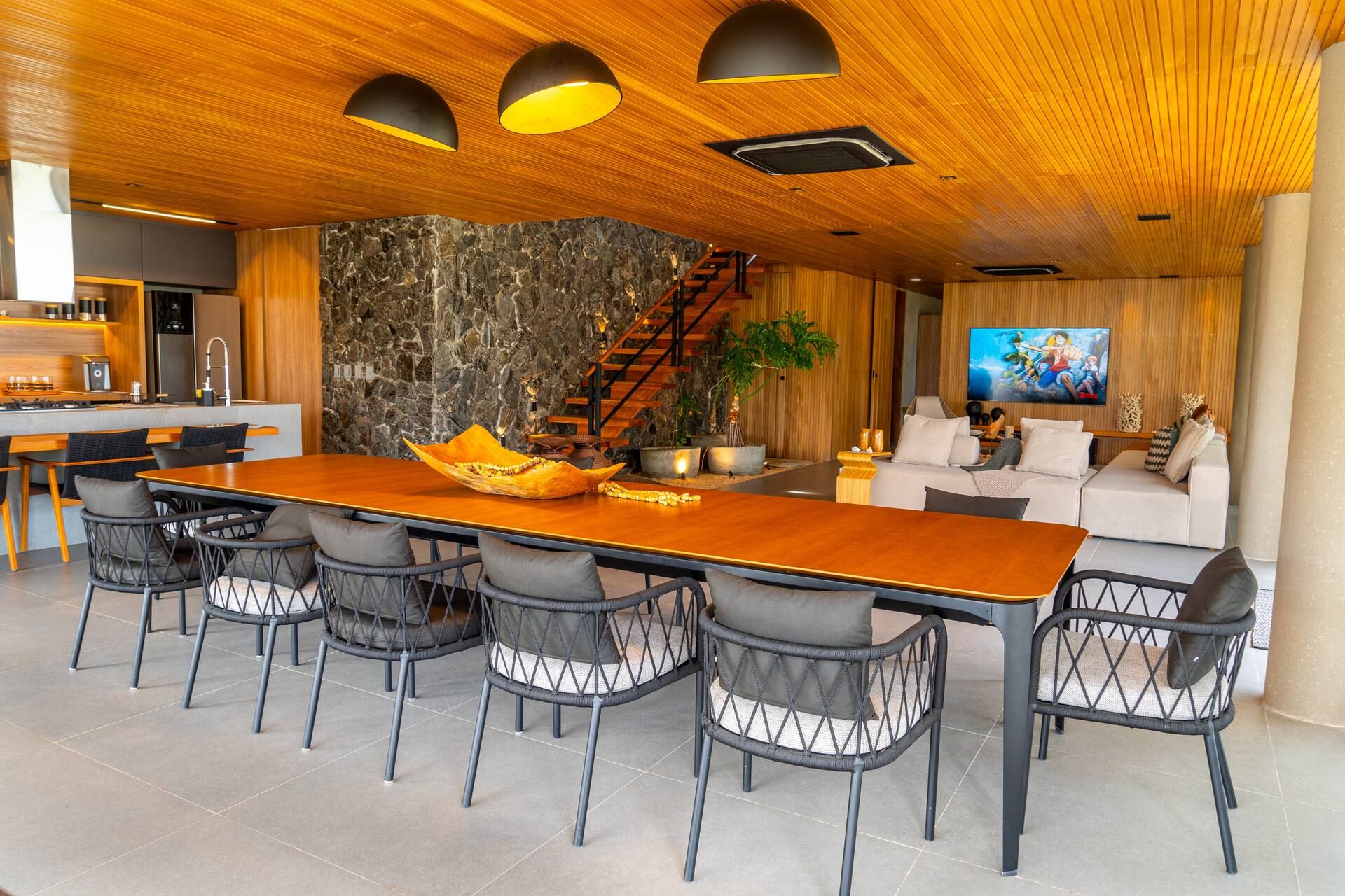 Sala de jantar climatizada com mesa para doze lugares, teto ripado de madeira e parede de pedra