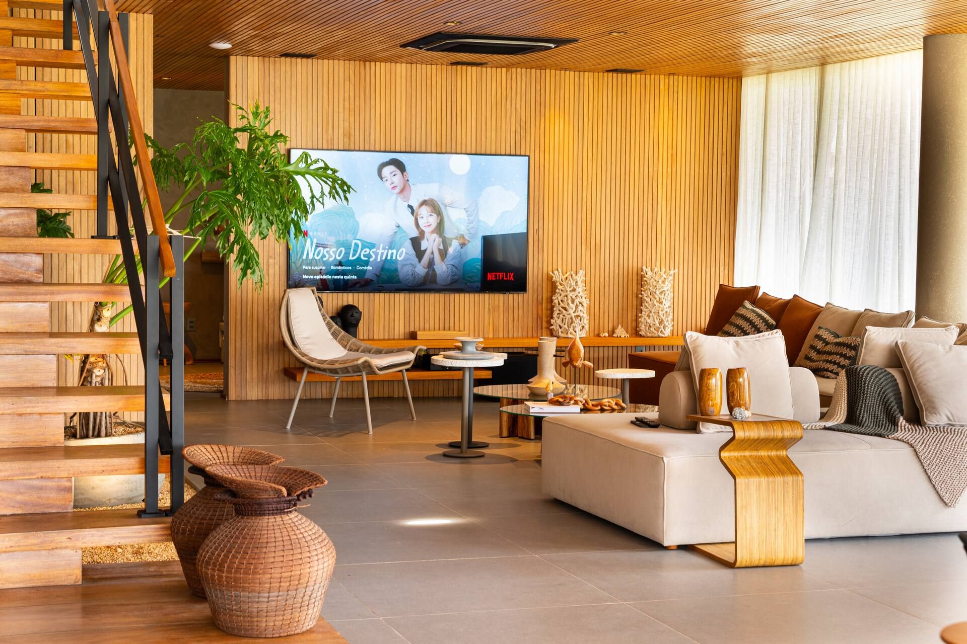Sala de estar da Casa Pérola com sofá claro, painel ripado de madeira, televisão e escada de acesso ao rooftop