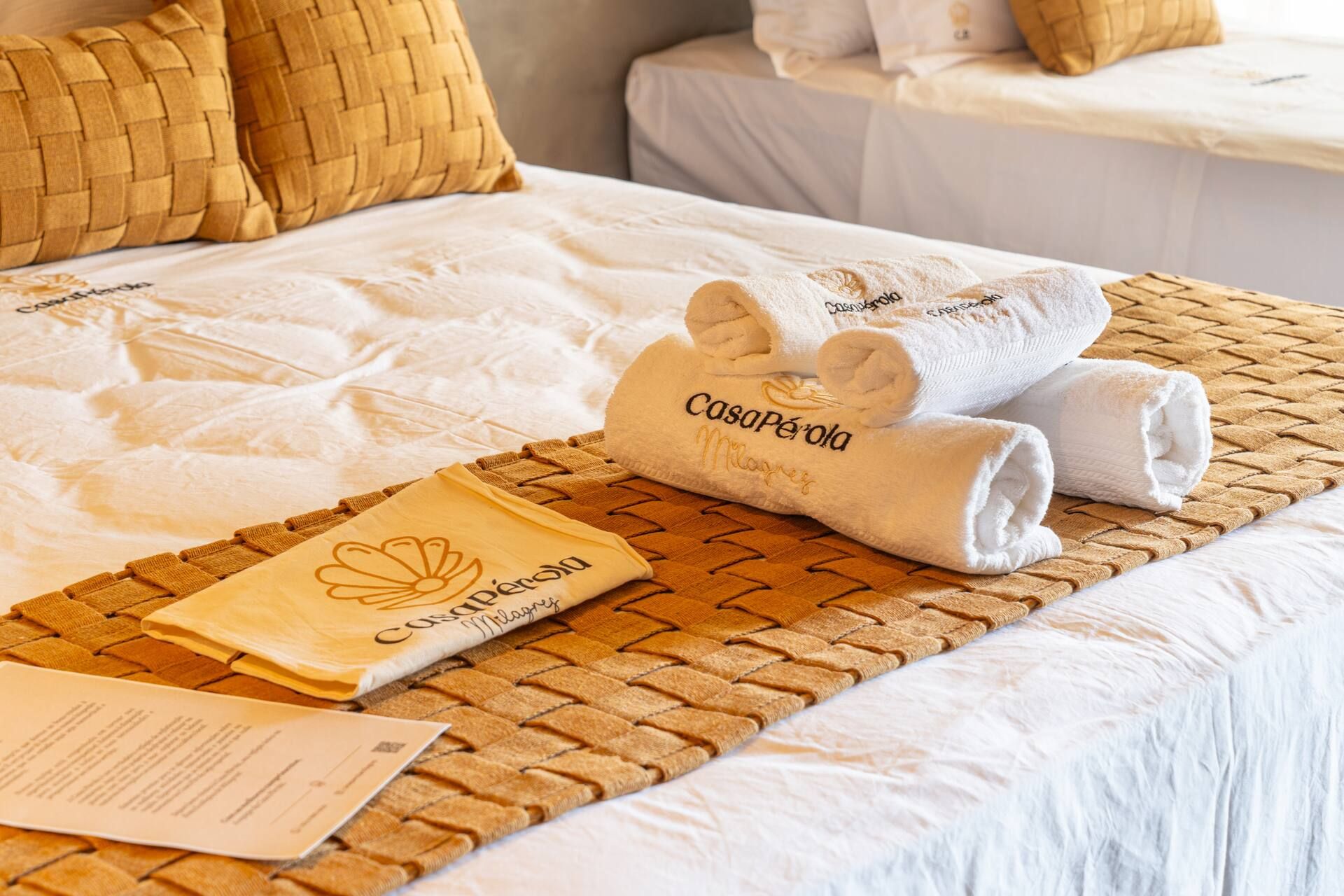 Enxoval da Casa Pérola sobre a cama, com toalhas brancas bordadas com o logotipo da casa