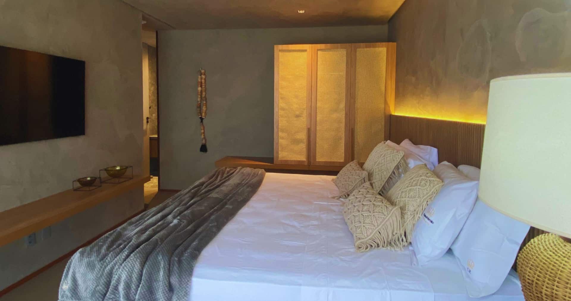 Suíte com painel ripado de madeira, televisão, luminária de piso e roupa de cama branca