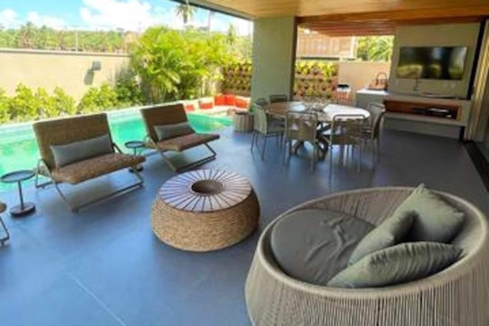 Lounge da varanda com poltronas de fibra natural, mesa de jantar externa e televisão
