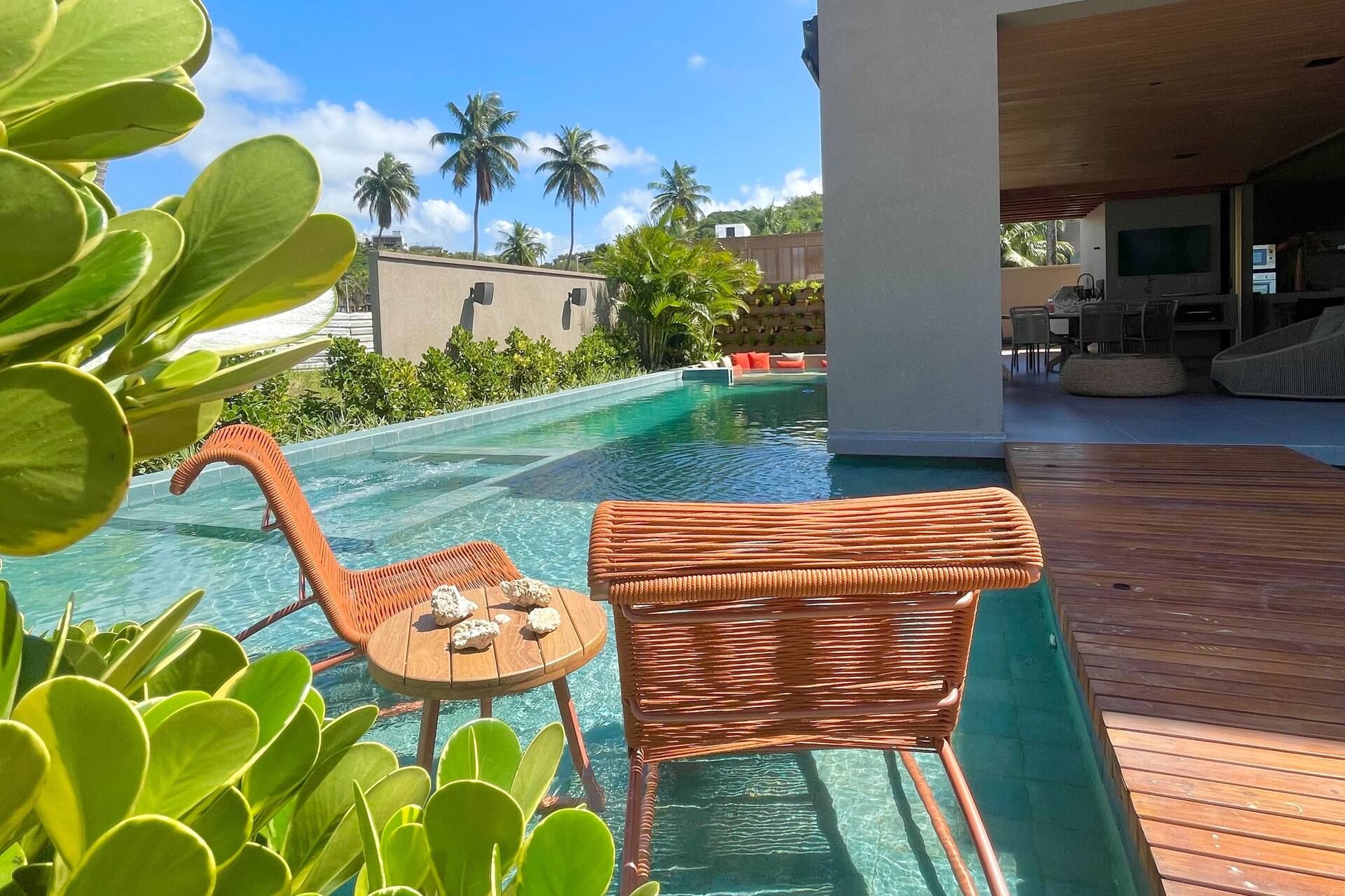 Borda da piscina com poltrona de vime e mesa de apoio, cercada por vegetação tropical